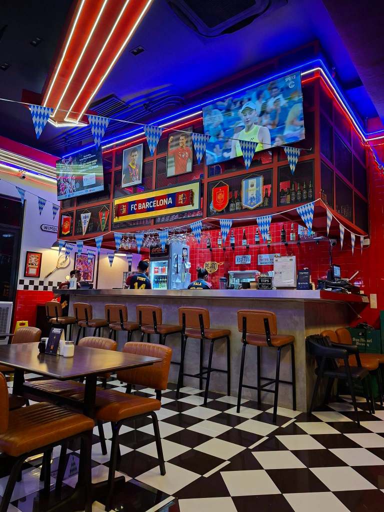 Interior of a sports-themed restaurant featuring a bar with FC Barcelona decorations, large television screens displaying sports events, and a checkered floor.