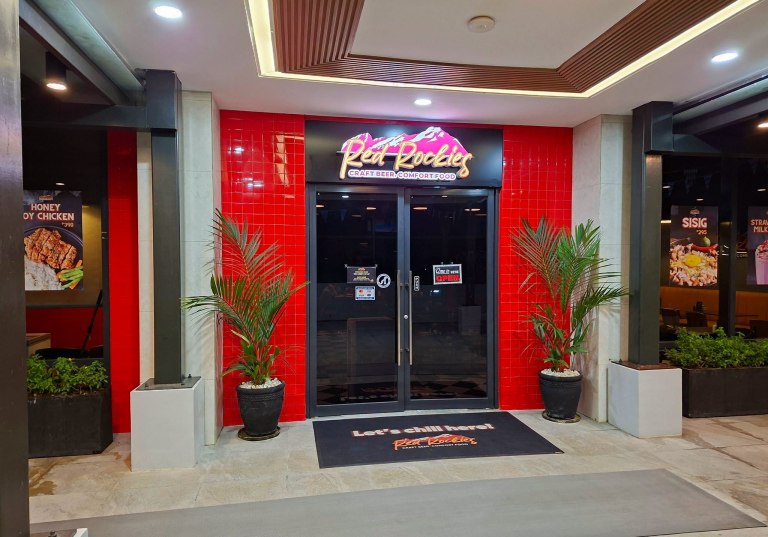 The entrance of 'Red Rookies', a restaurant featuring a bright red tiled facade, a glass door, and a sign that reads 'Craft Beer Comfort Food'. There are potted plants on either side of the entrance and a welcome mat that says 'Let's chill here!'