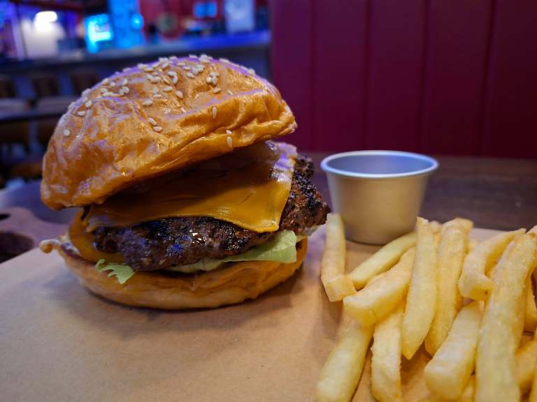 A close-up of a sesame seed burger with a beef patty and melted cheddar cheese, served with lettuce, accompanied by a small cup and a side of French fries.