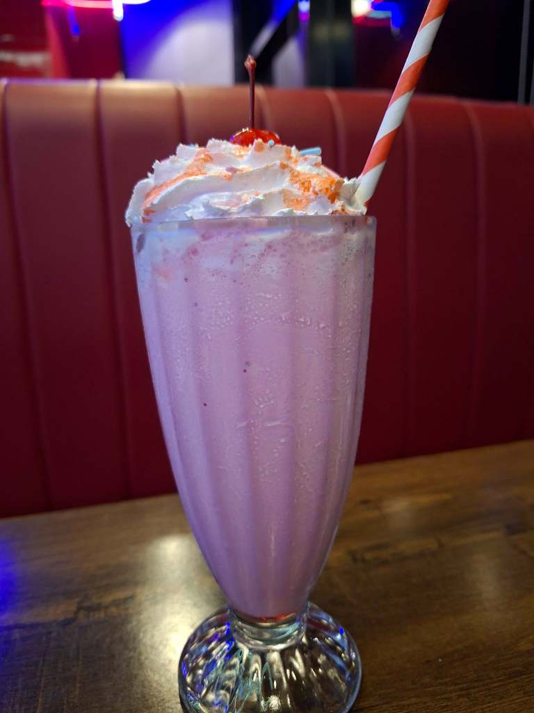 A tall glass of pink milkshake topped with whipped cream, colorful sprinkles, and a cherry, placed on a wooden table with a red upholstered background.